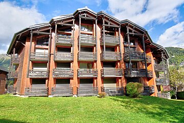 A multi-story wooden chalet-style building with many balconies sits on a green lawn, backed by mountains and a blue sky with clouds.