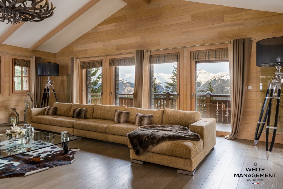 A cozy, wood-paneled chalet living room with a long sofa, fur throw, and panoramic views of snow-capped mountains through floor-to-ceiling windows.