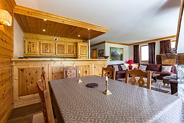 A cozy chalet interior with a patterned dining table, wood-paneled kitchen/bar, and a living room featuring sofas, a red lamp, and mountain art.