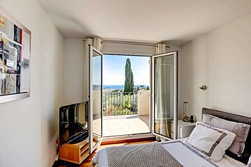A bright bedroom with open patio doors leading to a sunny balcony, offering a stunning sea, coast, and tree-lined view. A bed and TV are visible inside.