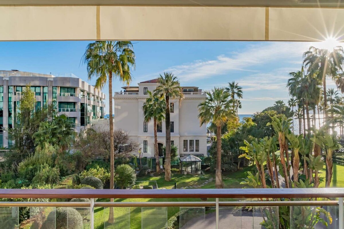 Balcony view of a sunny landscape with lush gardens, tall palm trees, diverse buildings, and the sparkling blue sea in the distance.