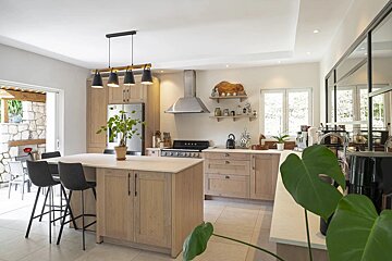 Bright, modern kitchen with light wood cabinets, a large island, stainless steel appliances, and plants. Open view to a stone patio.