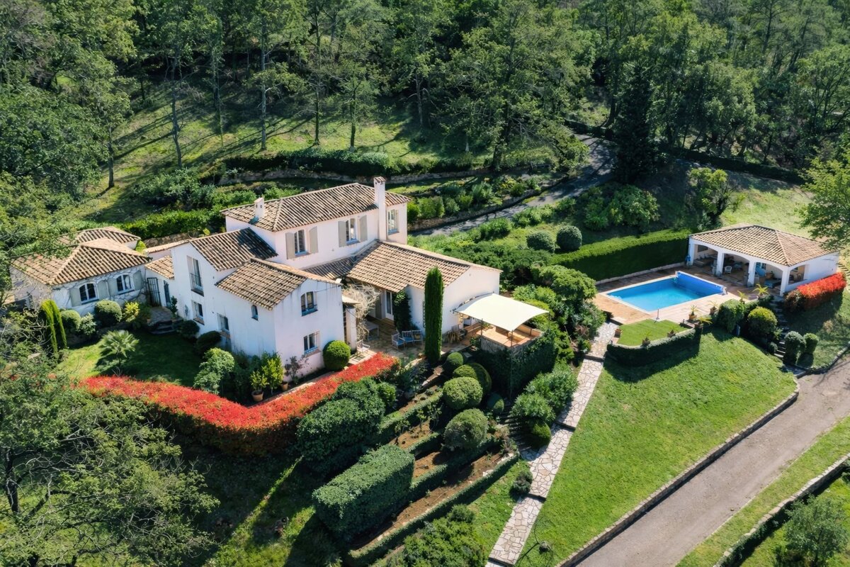 Aerial view of a white, tiled-roof French villa with manicured gardens, a pool, and a pool house surrounded by lush green hills.