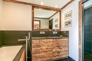 A rustic modern bathroom featuring a double wooden vanity, a large framed mirror, dark tile accents, and a bathtub.