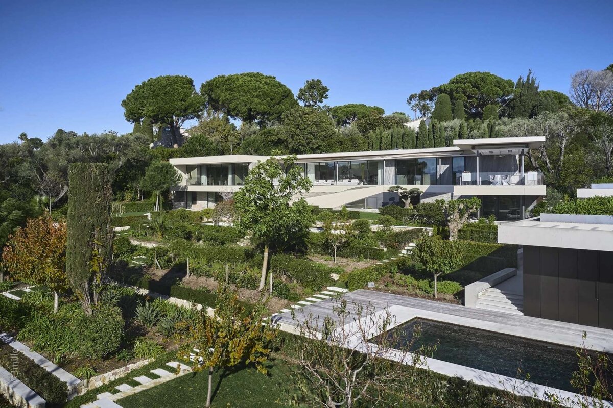 A modern hillside villa with extensive terraced gardens, a swimming pool, and lush trees under a clear blue sky.