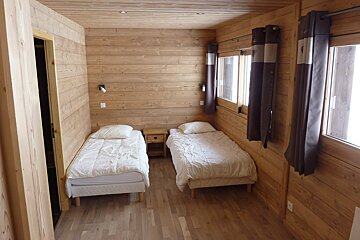 A cozy, wood-paneled room features two single beds with white bedding, a small nightstand, and windows draped with dark brown curtains.