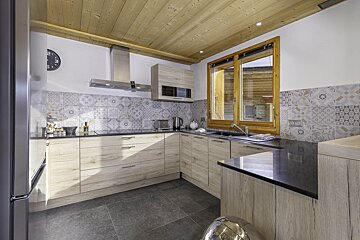 A modern kitchen with light wood cabinets, a wooden ceiling, patterned tile backsplash, and dark countertops.