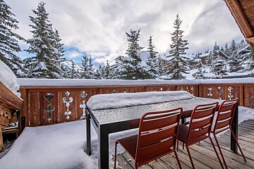 A snowy balcony with a table and red chairs, overlooking a winter landscape of snow-covered pine trees and distant homes under a cloudy sky.
