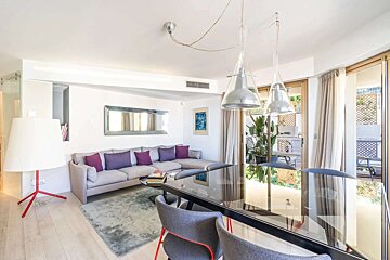 Stylish open-plan living/dining room. Features a grey sofa with purple cushions, black glass dining table, unique white lamp, and balcony access.
