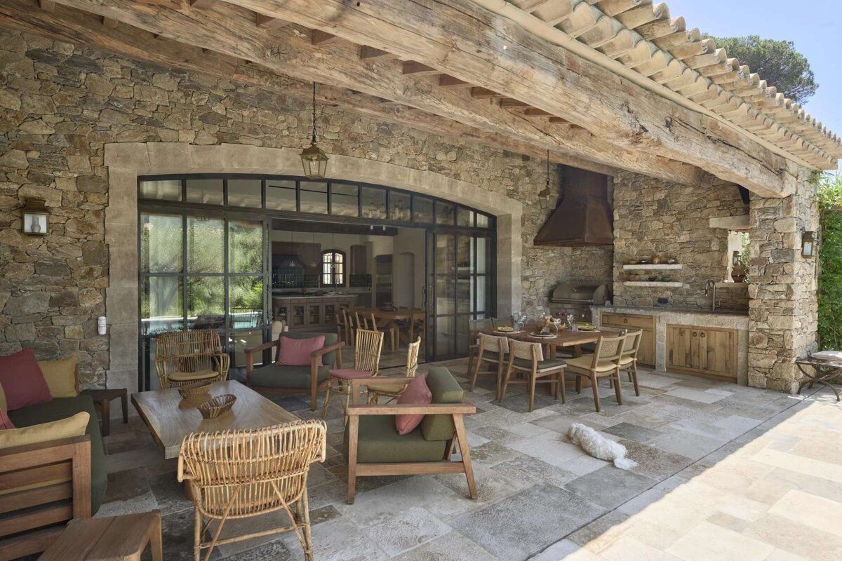 A rustic stone patio features outdoor living, dining, and kitchen areas under a beamed roof, with large glass doors and a white dog.