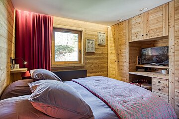 A cozy, wood-paneled chalet bedroom features a bed with lilac bedding, vibrant red curtains, a window, and a built-in entertainment unit with a TV.