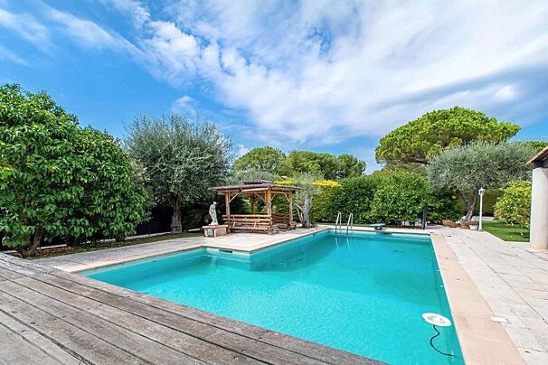 A refreshing blue swimming pool with a wooden deck and gazebo, nestled in a lush green garden under a bright blue, cloudy sky.