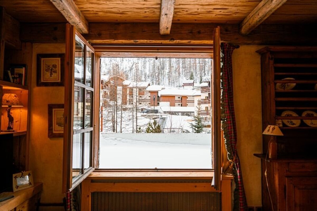 A cozy, rustic room with wooden beams looks out an open window to a snow-covered village and winter landscape.
