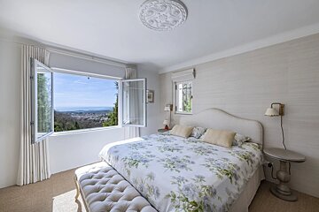 Bright bedroom with a floral bedspread and a large open window revealing a beautiful sea and landscape view.