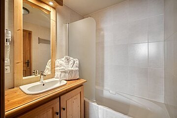 A modern rustic bathroom with a wooden vanity, large mirror, white sink, hairdryer, fluffy bathrobe, white tiled walls, and a bathtub.