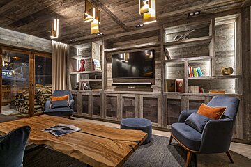 Rustic living room with built-in wooden shelving, a wall-mounted TV, two blue armchairs, and a live-edge wooden coffee table.