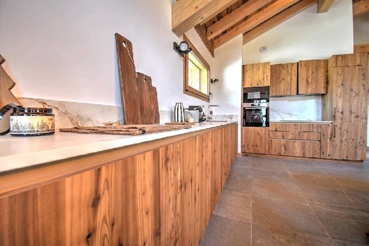 Rustic modern kitchen featuring warm natural wood cabinets, exposed ceiling beams, white countertops, and stone floor tiles.