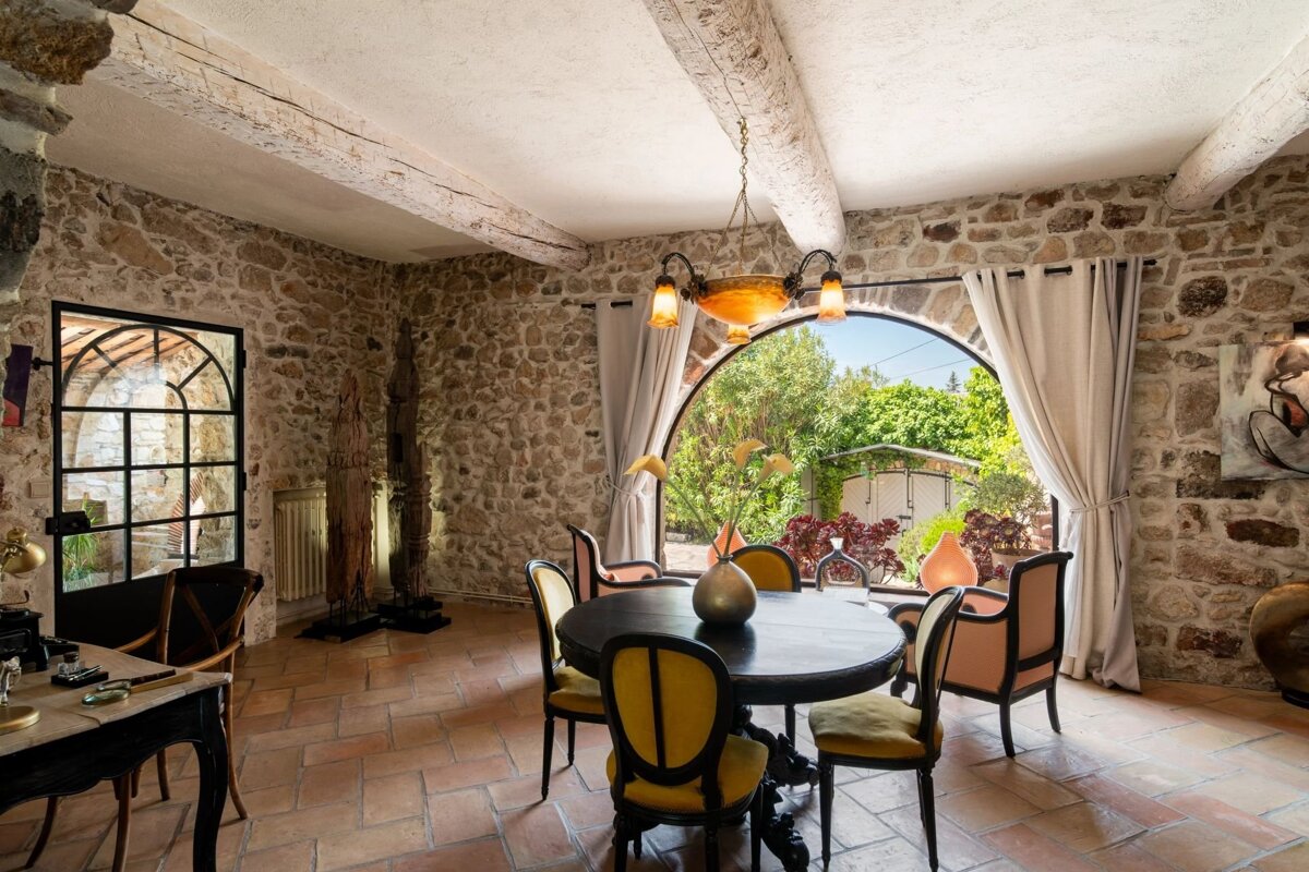 Rustic dining room with exposed stone walls, a round table with yellow chairs, and a large arched window overlooking a garden.