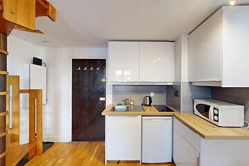 A modern kitchenette features white cabinets, light wood counters, a sink, stove, microwave, and mini-fridge. A dark door, hardwood floor, and wooden spiral staircase are also visible.