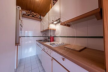A compact kitchen features white cabinets, wooden countertops and ceiling, white tiled walls with a green stripe, a sink, stove, and a wall-mounted wine rack.
