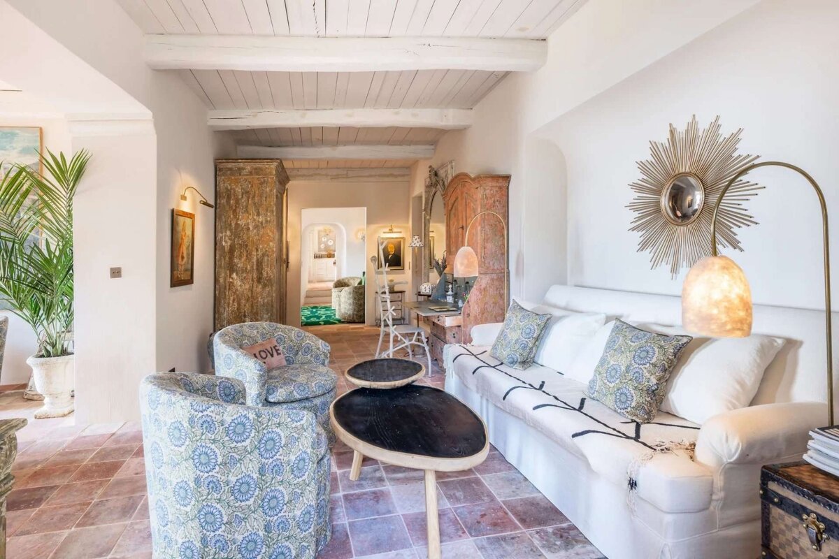 A bright, rustic living room with exposed beams, terracotta floor, white slipcover sofa, patterned chairs, antique wooden cabinets, and a sunburst mirror.
