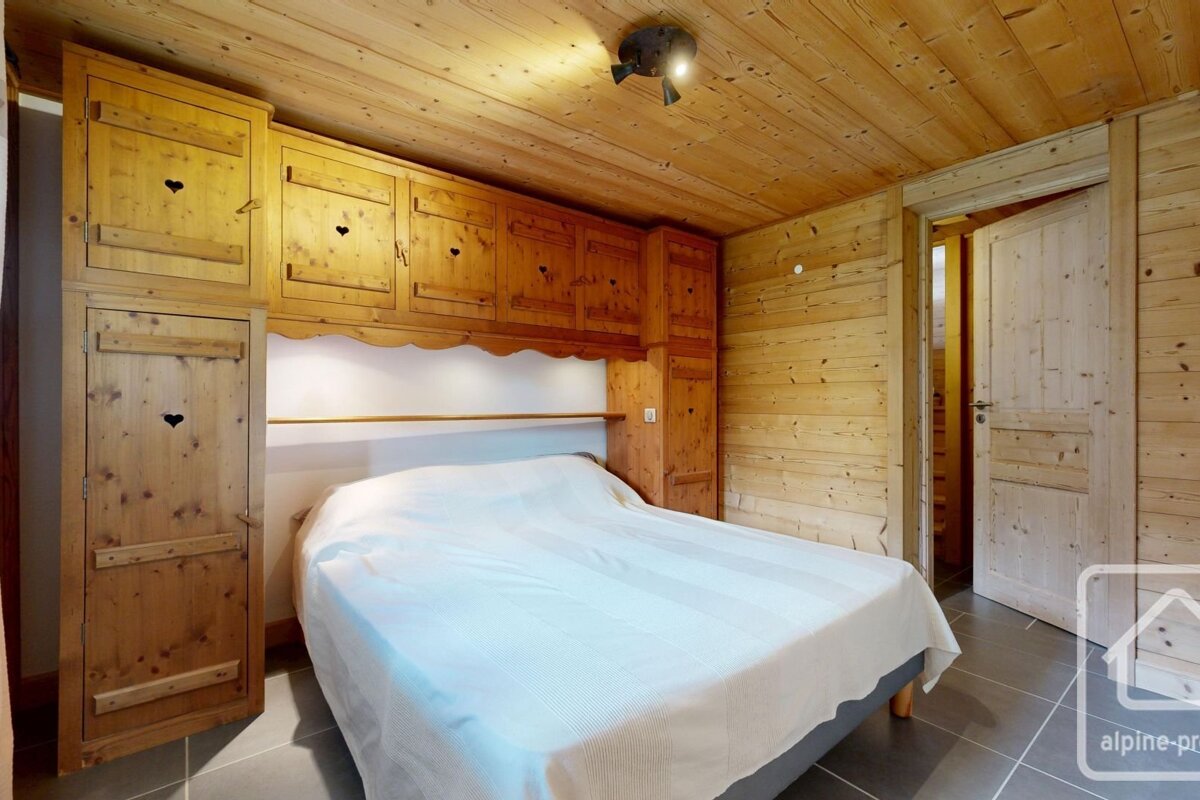 Cozy, rustic bedroom with wooden walls, ceiling, and built-in storage featuring heart cutouts. A bed with a white cover is centered.