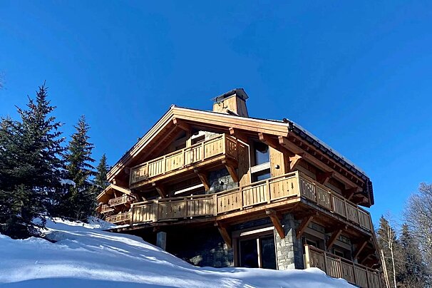 Modern wooden chalet on a snowy mountain. Stone base, multiple balconies, and pine trees under a clear blue sky.