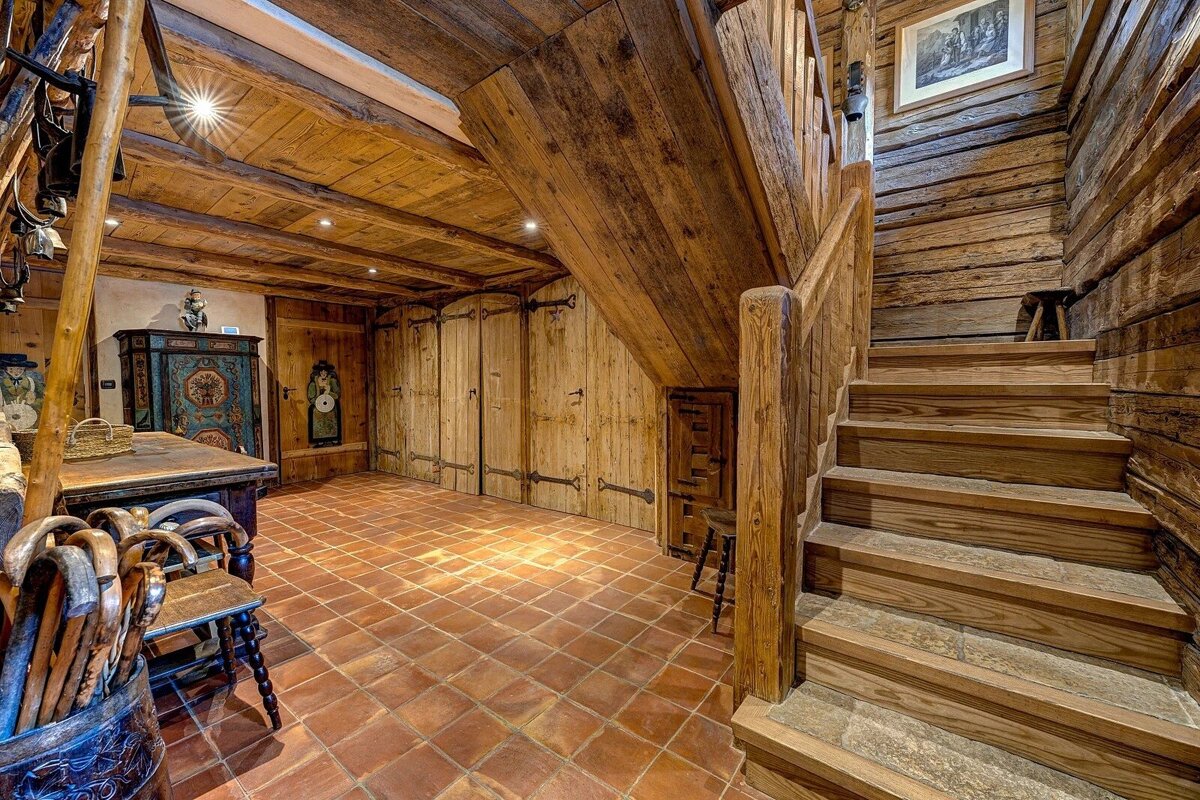 A cozy, rustic wooden interior featuring a prominent staircase, timber walls and ceiling, a terracotta tiled floor, and antique furniture.
