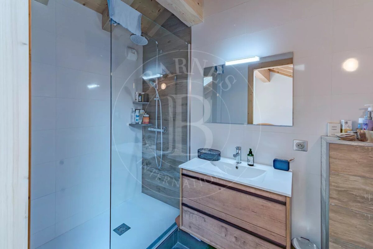 A bright, modern bathroom features a glass shower, wooden vanity with sink and mirror, white tiled walls, and wooden ceiling beams.