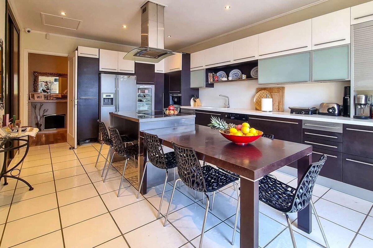 A modern kitchen with dark and white cabinets, stainless steel appliances, an island, and a dining table with chairs. Fruit bowls add color. Tiled floor.
