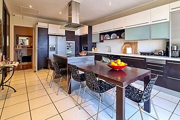 A modern kitchen with dark and white cabinets, stainless steel appliances, an island, and a dining table with chairs. Fruit bowls add color. Tiled floor.