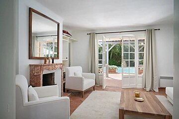 Bright white living room featuring a marble fireplace, cozy armchairs, and French doors that open to a sunny pool patio.