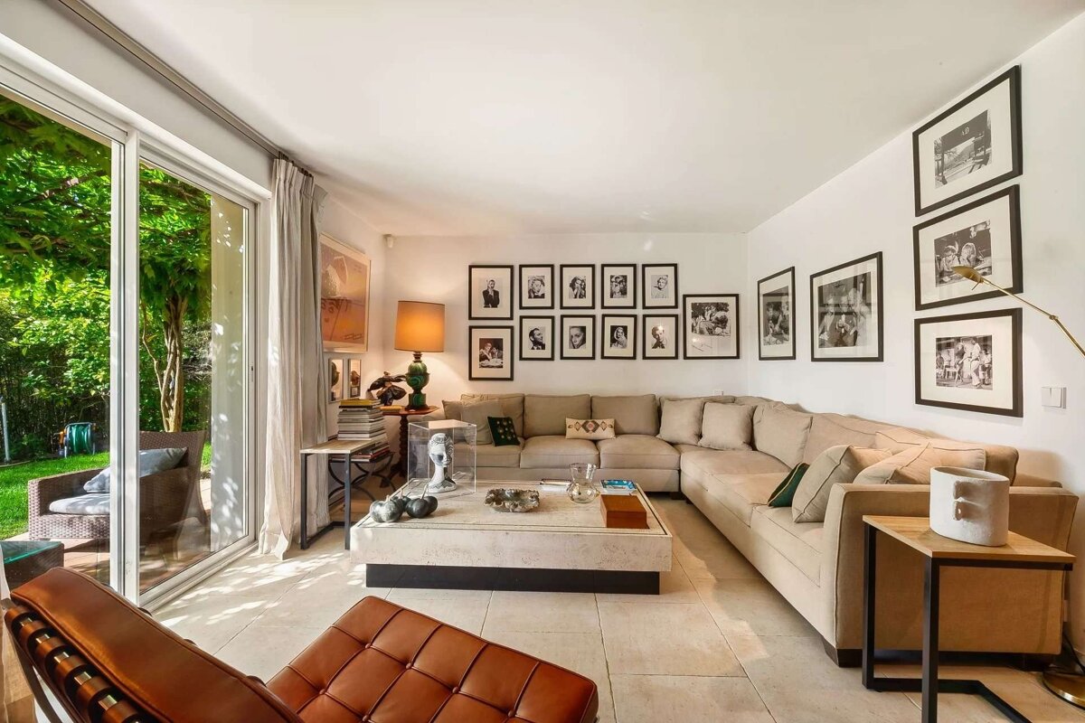 A bright, spacious living room features a large beige sectional sofa, gallery wall of photos, and glass doors opening to a lush green garden.