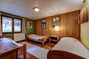 Rustic bedroom with three single beds, wood paneling, a green wall, and a window view. Wooden furniture and white bedding create a cozy feel.