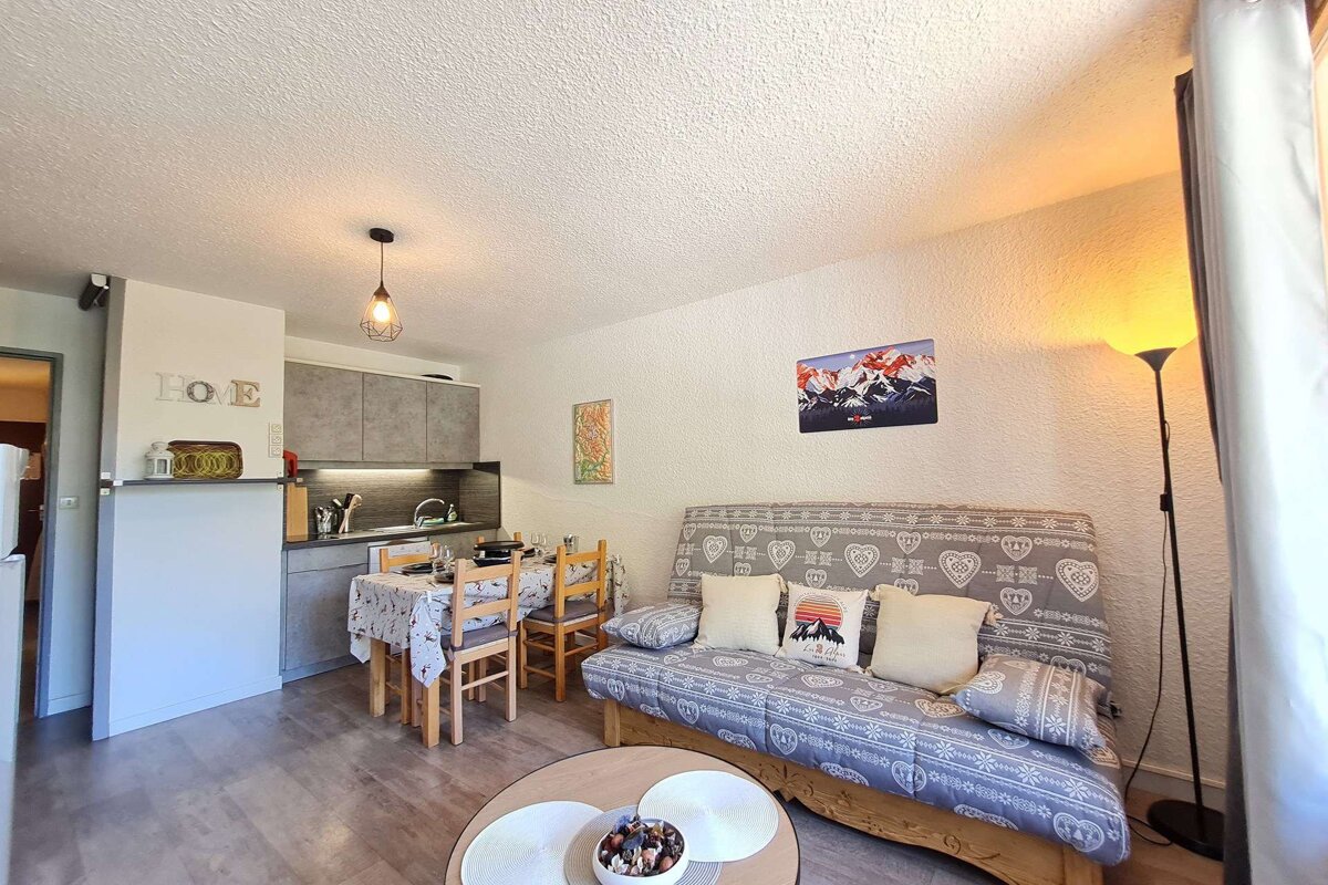 A cozy open-plan living space features a modern grey kitchen, a dining table, and a patterned sofa with mountain-themed decor. Warm and inviting.