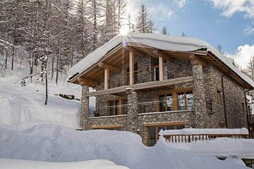 A rustic stone chalet covered in deep snow, set against a winter forest with bright sun flaring over the rooftop.