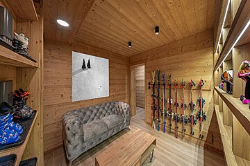 A warm, wood-paneled ski chalet interior featuring organized ski gear, a comfortable grey sofa, and a minimalist snowy landscape artwork.