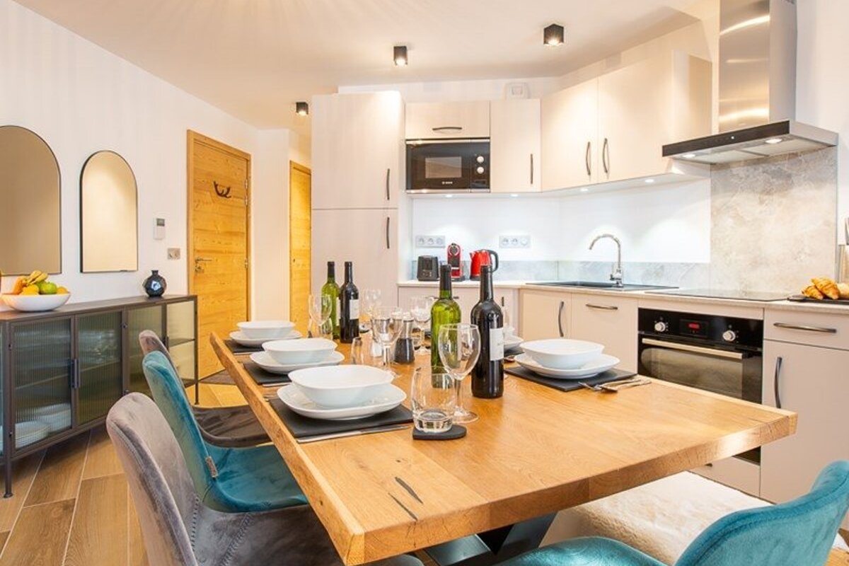 Modern open-plan kitchen and dining area featuring a wooden table set for a meal, elegant velvet chairs, and sleek white cabinets.