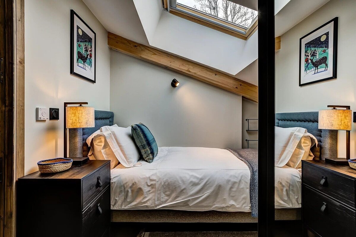 Cozy attic bedroom with a skylight, wooden ceiling beam, and a mirror reflecting the bed and nightstand.