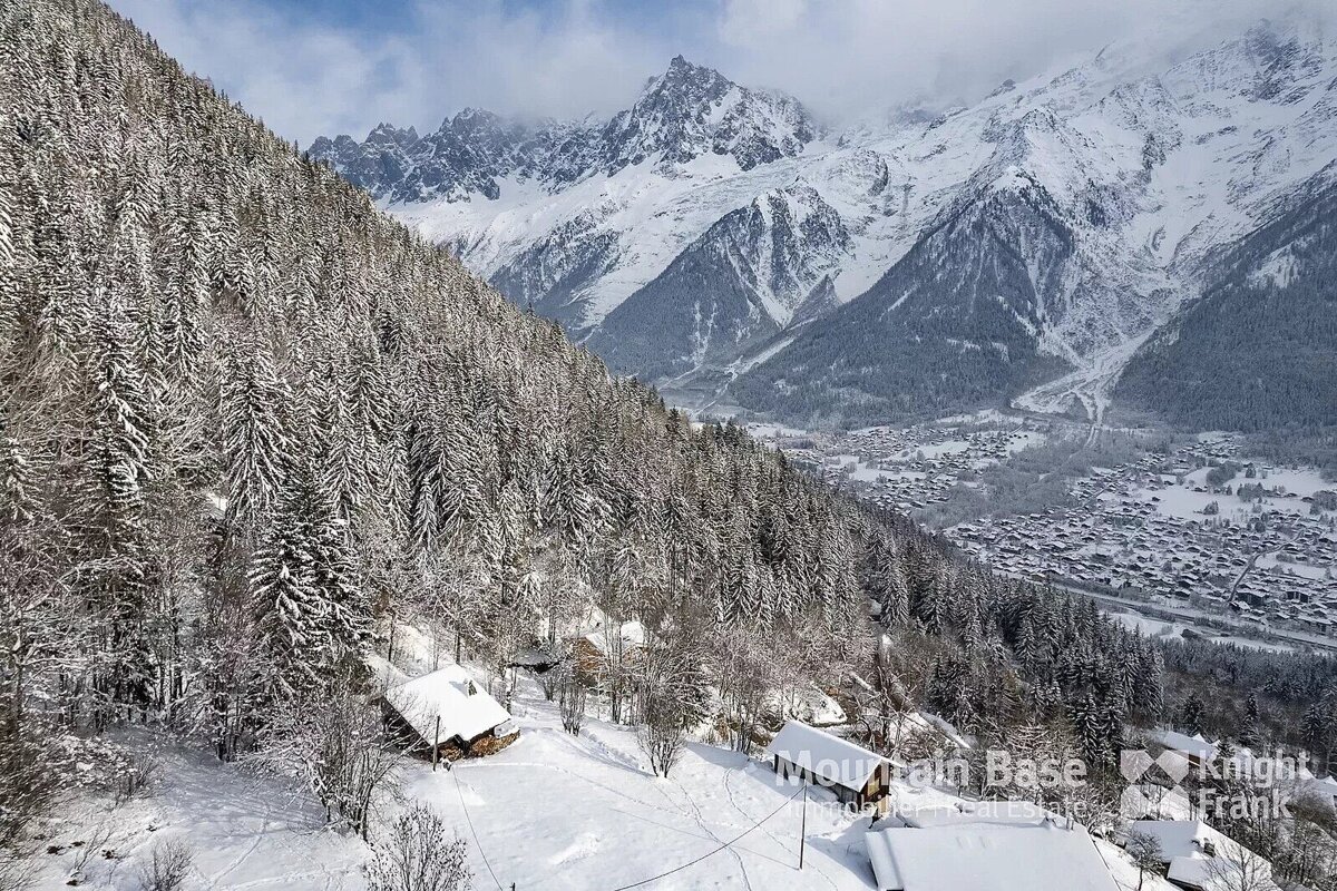 A snow-covered mountain slope with dense pine forests and chalets, overlooking a distant valley town nestled among majestic, snow-capped peaks.