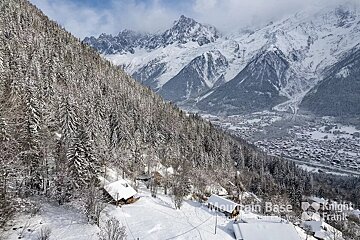 A snow-covered mountain slope with dense pine forests and chalets, overlooking a distant valley town nestled among majestic, snow-capped peaks.