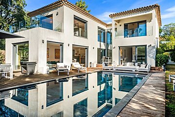 Modern white villa with large glass windows, a sparkling pool reflecting the house, and a spacious wooden deck on a sunny day.