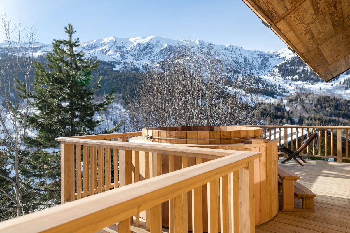 A wooden hot tub sits on a sunny deck, offering a stunning view of majestic snow-capped mountains, evergreen trees, and a clear blue sky.