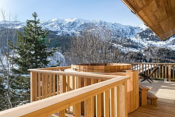 A wooden hot tub sits on a sunny deck, offering a stunning view of majestic snow-capped mountains, evergreen trees, and a clear blue sky.