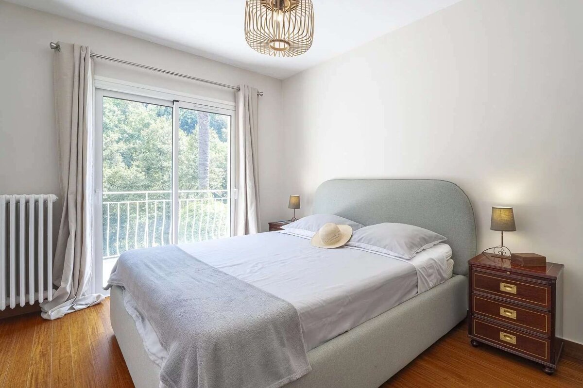 Bright, modern bedroom with a light gray bed, straw hat, dark wood nightstands, and a glass door opening to a view of green trees.