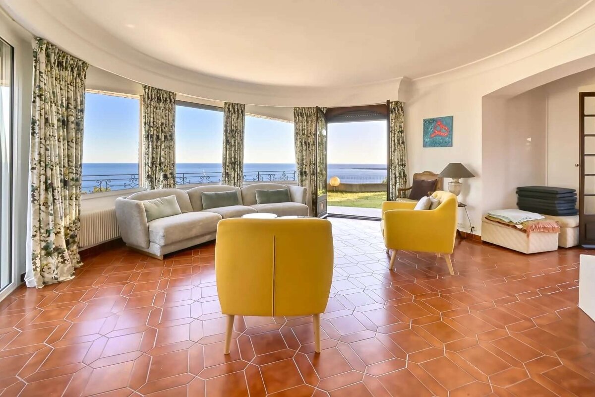 Spacious living room with curved windows overlooking the sea, featuring a light sofa, bright yellow armchairs, and terracotta geometric tiled floor.