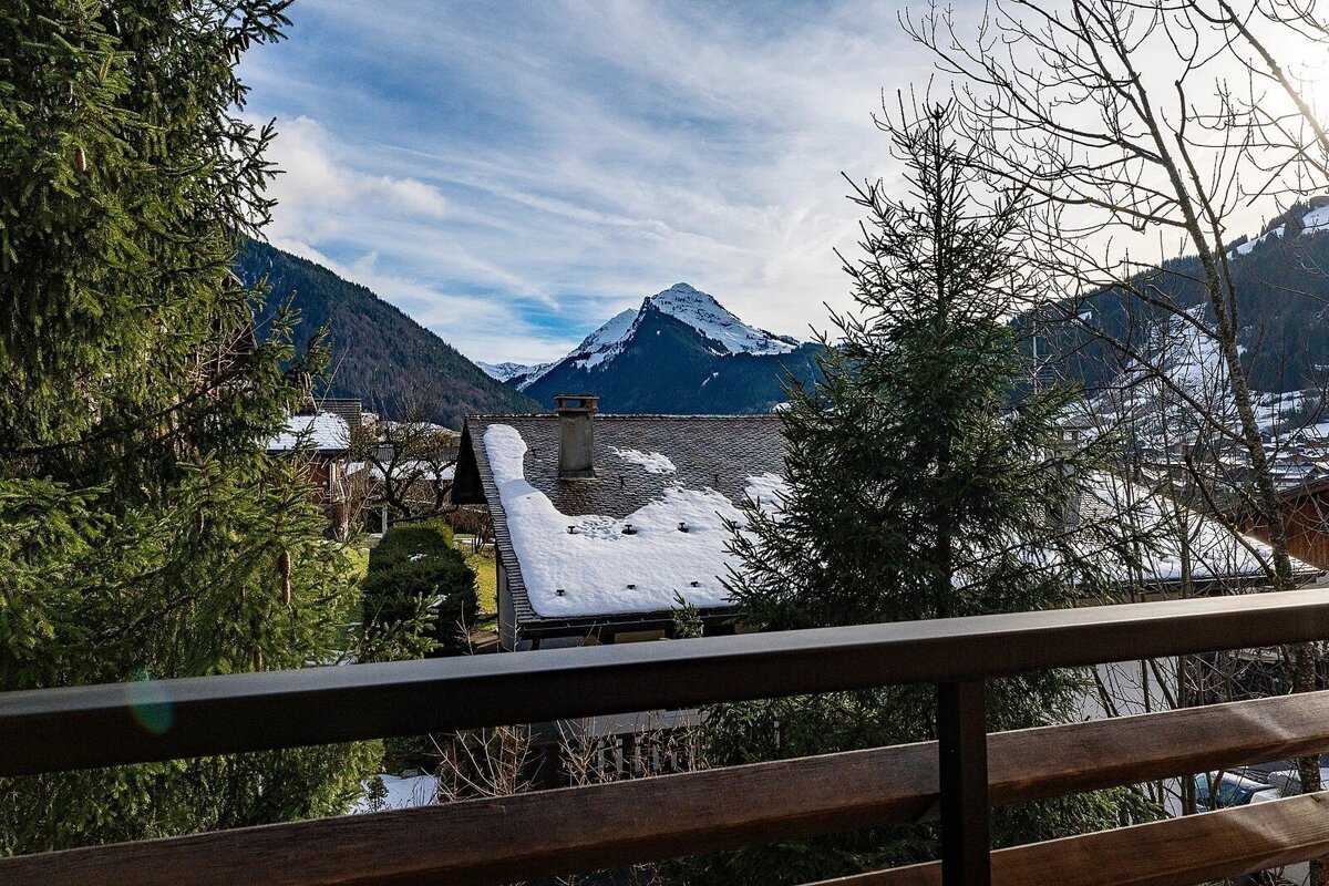A balcony view of a snowy alpine village with houses and evergreen trees, framed by a majestic, snow-capped mountain peak under a partly cloudy blue sky.