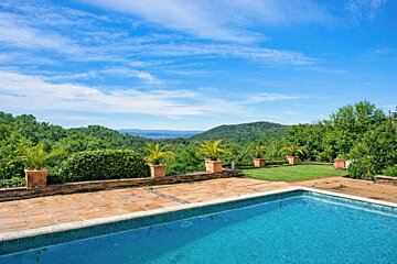 A refreshing blue pool and tiled patio overlook lush green hills stretching to the horizon under a bright blue, cloud-streaked sky.