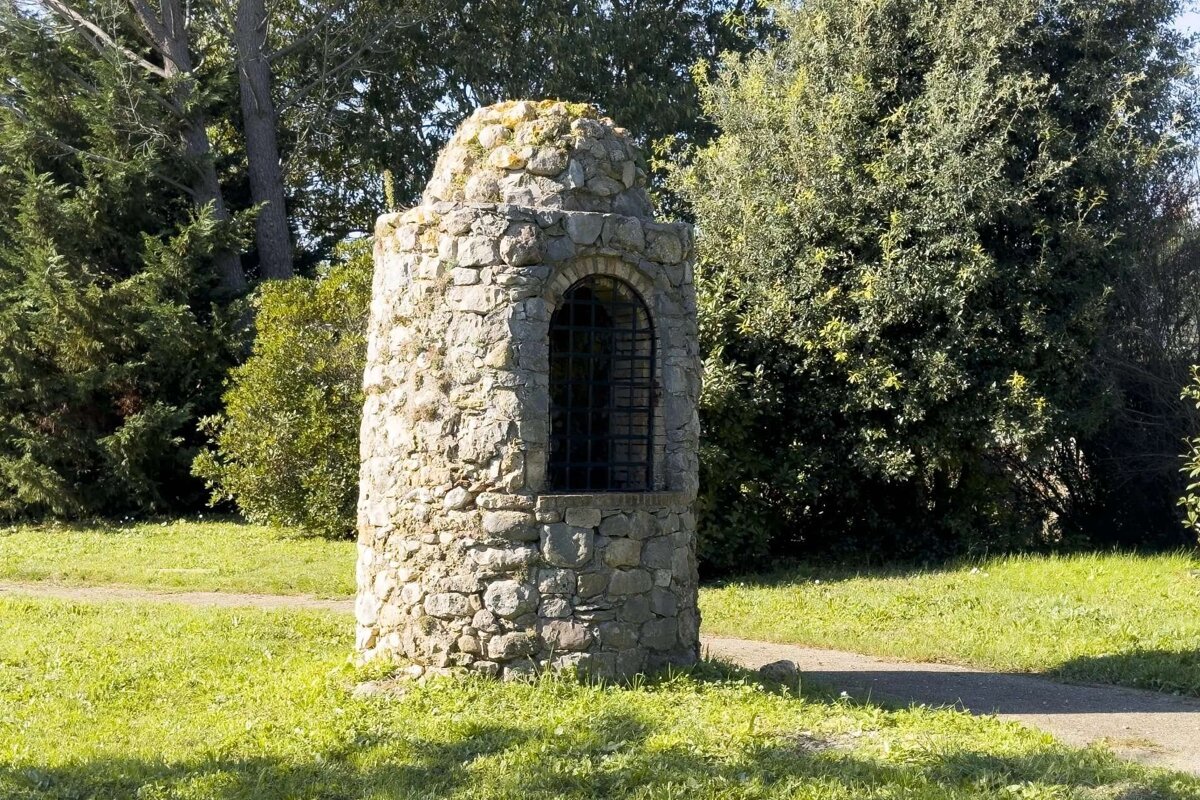 A small stone tower with an arched, barred window and mossy rounded top stands in a sunny, grassy park surrounded by green trees.