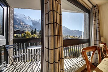A bright room with checkered curtains and wooden chairs opens to a balcony. It offers a sunny view of a forested valley and snow-capped mountains.